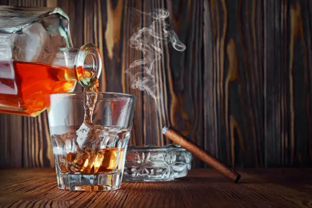 Strong alcoholic drink scotch whisky pour from decanter in old fashion glass with ice cube and smoking cigar in ashtray on wood texture background