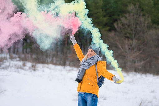 Beautiful, young woman having fun with colorful smoke bombs on the snowy mountain.