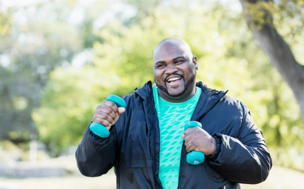 Mature African-American man exercising A mature African-American man in his 40s with shaved head, mustache and beard, exercising in the park with hand weights, smiling at the camera. He has a large build. overweight man stock pictures, royalty-free photos & images