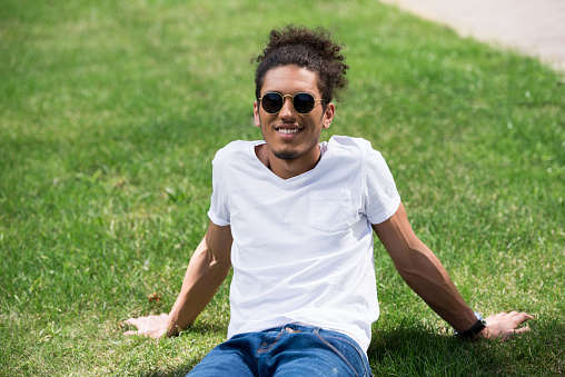 handsome young african american man in sunglasses sitting on grass and smiling at camera