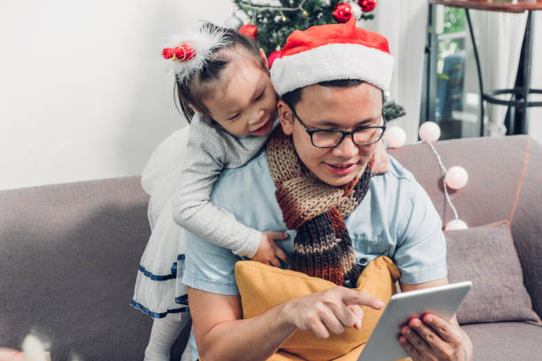 father and daughter sitting on sofa  and select christmas present and shopping from online store by tablet at xmas party,holiday gift giving festive. - father digital tablet asian ethnicity daughter imagens e fotografias de stock