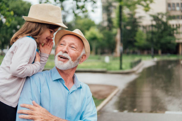 ragazza che sussurra al nonno al parco - whispering grandparent child grandfather foto e immagini stock