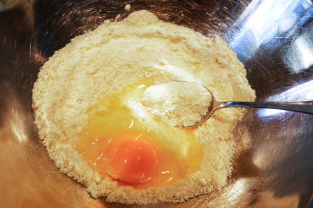 glutenfrei kochen low-carb - ein ei in einen haufen von mandel-mehl mit einem löffel bereit, es in eine rostfreie schüren gebrochen stehlen schüssel - nahaufnahme - low back stock-fotos und bilder