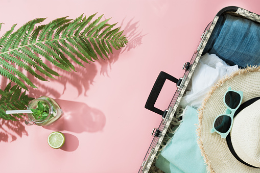 Leaf of fern, tropical detox water and open suitcase with clothes on pastel pink background. Top view with copy space. Summer concept travel.