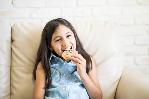 Young cute latin girl enjoying healthy cookie sitting on couch at home