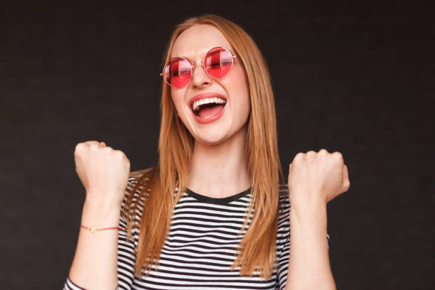 Attractive young woman in stylish sunglasses screaming and gesturing with hands while rejoicing over victory on black background
