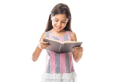 Little smart girl holding a book and reading it while standing against white background