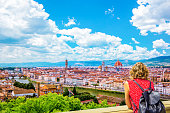 Woman tourist in red admires Florence Firenze (Duomo, Arno River, towers, cathedrals, tiled roofs of houses) from Piazzale Michelangelo, cityscape panorama top view, Florence, Tuscany, Italy