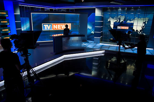 TV news studio ready for filming. Tv news anchor sitting in his place and two camera men silhouettes in front of him.