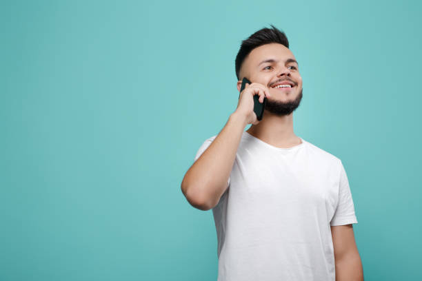 istock Handsome young bearded hipster in white t-shirt using phone and talking with bright smile. Copy space. 991248920