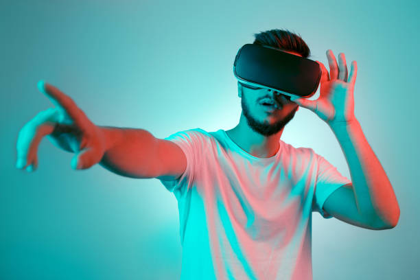 istock Handsome bearded man trying VR headset and poniting. Young man exploring another world with virtual reality goggles on the colorful background. 991249662