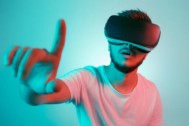 istock Attractive bearded man trying VR headset and poniting. Young man exploring another world with virtual reality goggles on the colorful background. 991250604