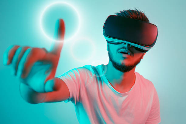istock Attractive bearded man trying VR headset and poniting in lightning circle. Young man exploring another world with virtual reality goggles on the colorful background. 991250680