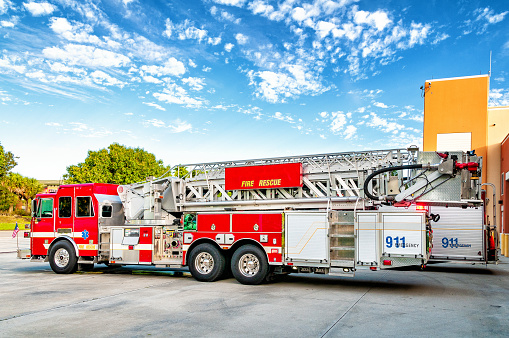 A hook and ladder fire engine under blue skies sits in front of the fire station.