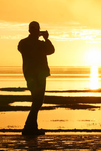 istock Silhouetted man back-lit by setting sun taking photo with mobile device 991366056