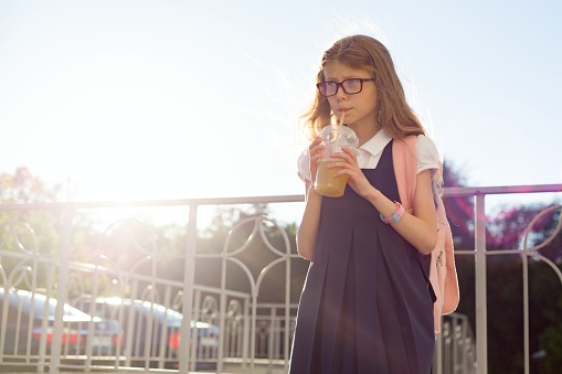 Outdoor portrait of girl elementary school student wearing glasses, school uniform, with backpack drinking natural juice from glass. Copy space