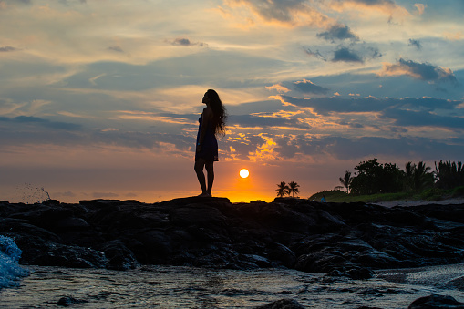 Silhouette of woman standing on a rocky Hawaii coastline enjoying the Hawaiian Sunset