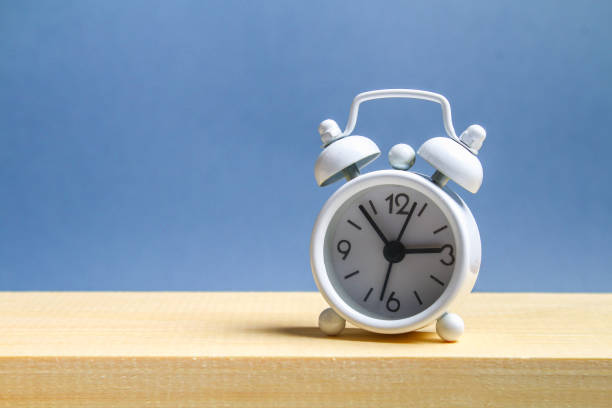 istock White small alarm clock on a wooden shelf on a grey pastel background. Minimalism. 991439792