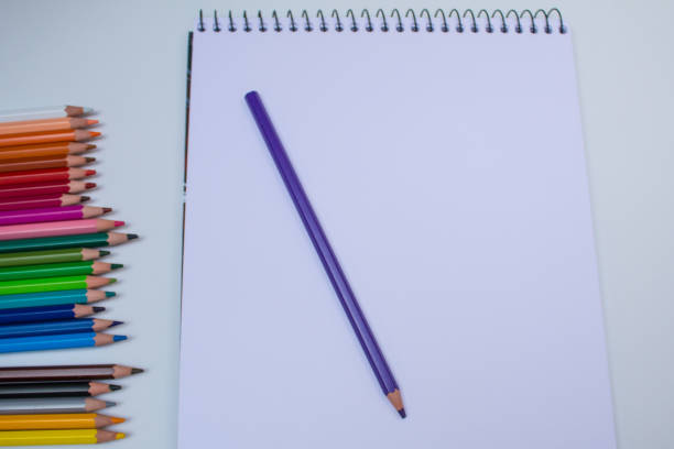 istock Drawing notebook with blue pencil on white background and colored pencils on left side 991527032