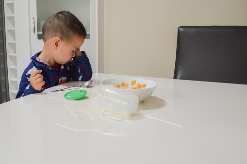 A candid photograph of a 2-year old Eurasian boy having breakfast in his pyjamas, and upset over spilling his cup of milk on the table