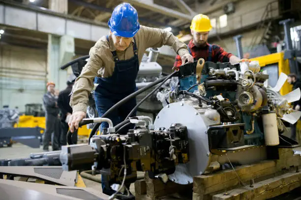 Photo of Concentrated skilled engineers in hardhats assembling tractor engine while attaching hose together in industrial workshop