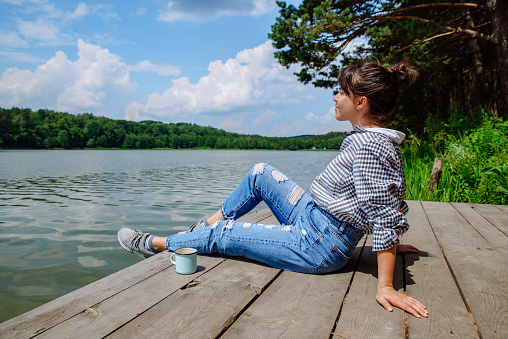 young adult woman sitting on wooden dock drinking coffee and looking at lake