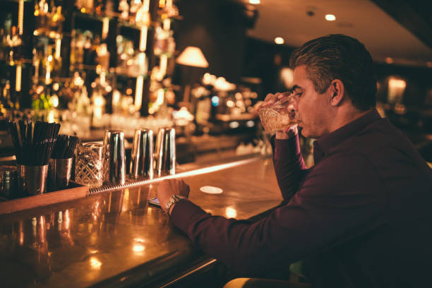 Elegant mature man at bar drinking and checking the time Elegant senior man sitting on bar counter drinking whiskey and looking at watch vex nightclub stock pictures, royalty-free photos & images