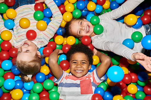 Above view portrait of three happy little kids in ball pit smiling at camera raising hands while having fun in children play center, shot with flash