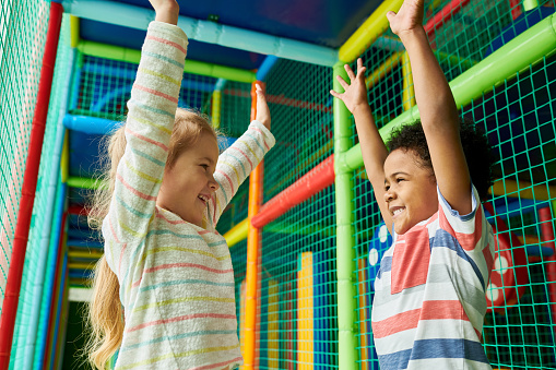 Colorful waist up portrait of happy boy and girl facing each other and raising hands in excitement while having fun in children play area