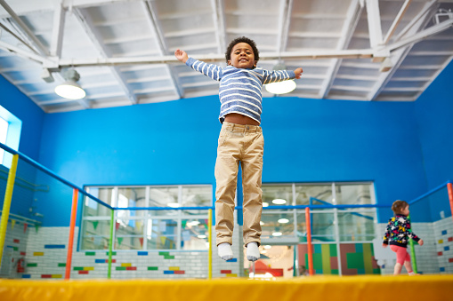 Full length low angle portrait of happy African-American boy jumping on trampoline in colorful kids play center, copy space