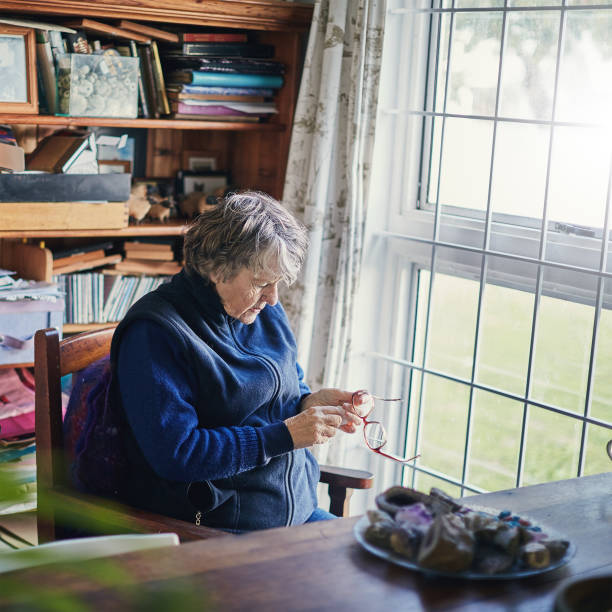 istock Senior woman sitting by window cleans her spectacles 991851574