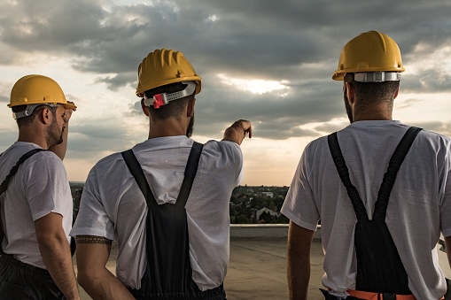 Rear view of construction workers standing on a roof while one of them is aiming at distance.