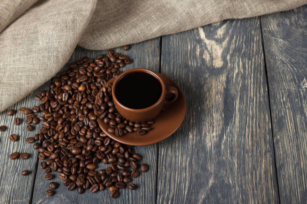 istock Cup of coffee, scattered grains on wooden surface, next to burlap 991873470