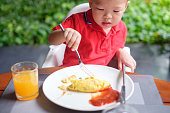 かわいい小さなアジア 2 年古い幼児赤ちゃん男の子赤シャツを着て使用フォークとナイフの庭で朝食を食べる