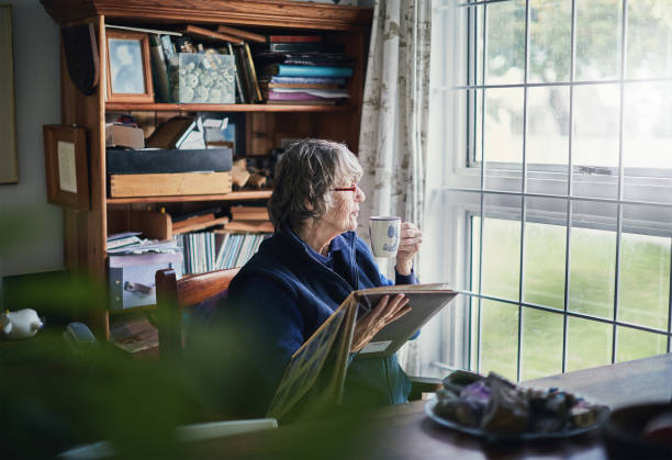 Senior woman holding photo album sips tea, looking through window An old woman holding a family photo album looks out through the window, lost in her memories. solitude photos stock pictures, royalty-free photos & images