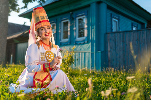 Adorable young woman dressed in women's traditional clothes and jewelry which are recreated according to medieval samples of the Mari people. The young woman is sitting on lawn next to an old village house. The young woman is smiling looking at the camera. Shooting in summer at sunset, Republic of Mari El in Russia's Volga region