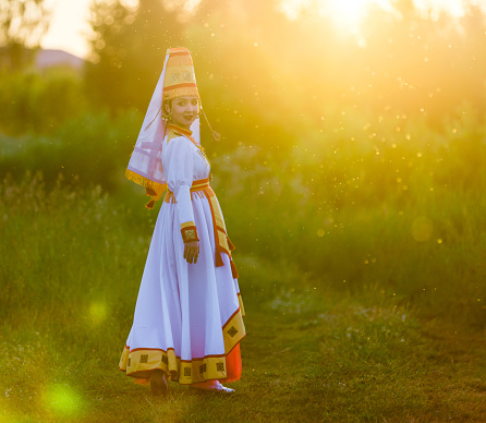 Adorable young woman dressed in women's traditional clothes and jewelry which are recreated according to medieval samples of the Mari people. The young woman is walking on a meadow on backgdrop a sun rays. The young woman is smiling looking over shoulder at the camera. Shooting in summer evening, Republic of Mari El in Russia's Volga region