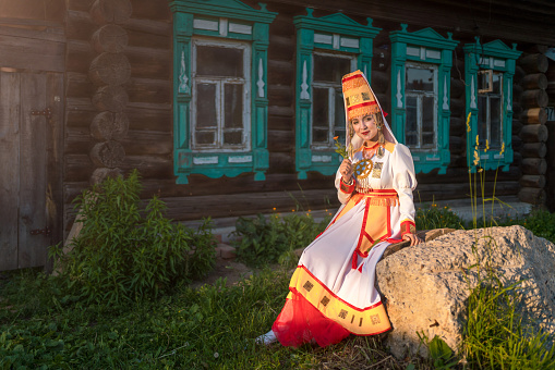 Cute young woman dressed in women's traditional clothes and jewelry which are recreated according to medieval samples of the Mari people. The young woman sits on a stone before the old country cottage. She is smiling looking at the camera. Shooting in a countryside at summer sunset, Republic of Mari El in Russia's Volga region
