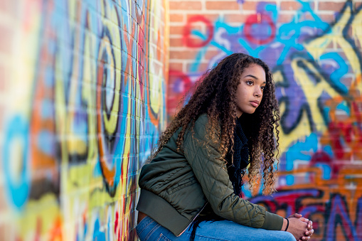 A high school student of African descent is sitting near a wall with graffiti on it. She is looking into the distance.