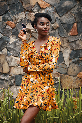 Slim African-American woman wearing summer floral dress and holding sunglasses while standing outside looking away