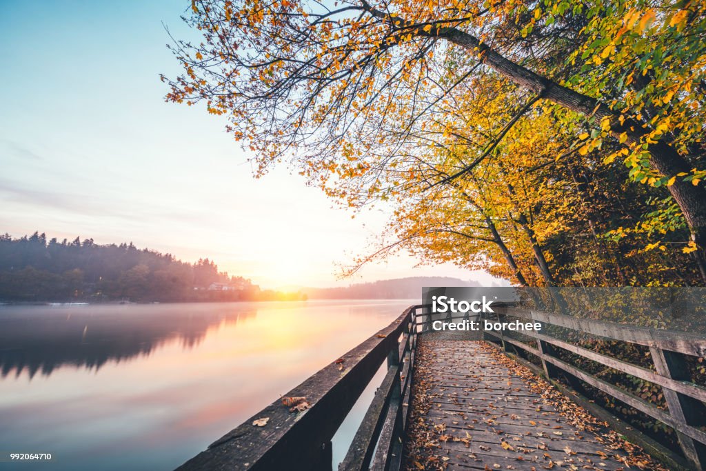 Sunrise By The Lake Footpath with a wooden fence by the misty lake. Early morning view with sun shinning through the fog. Autumn Stock Photo