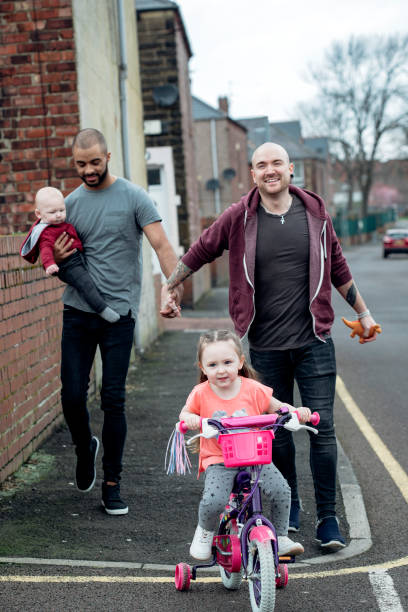 Male Couple Out Walking With The Family Gay male couple taking a walk with their baby boy and holding hands. Their young daughter rides her bicycle down the street in front of them. skin head stock pictures, royalty-free photos & images