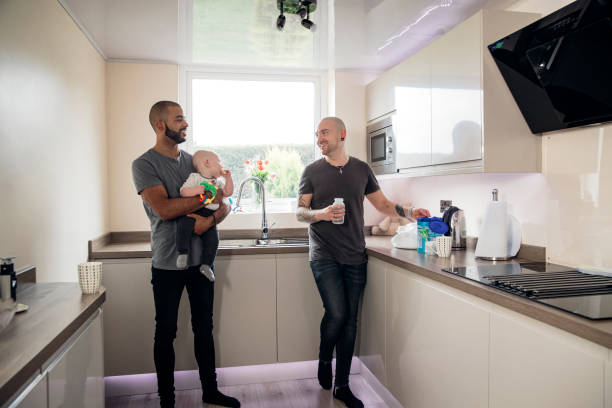 Male Couple at Home Couple spending time with their little baby boy. They're all together at home standing in the kitchen. One man is holding a baby boy gay daddy stock pictures, royalty-free photos & images