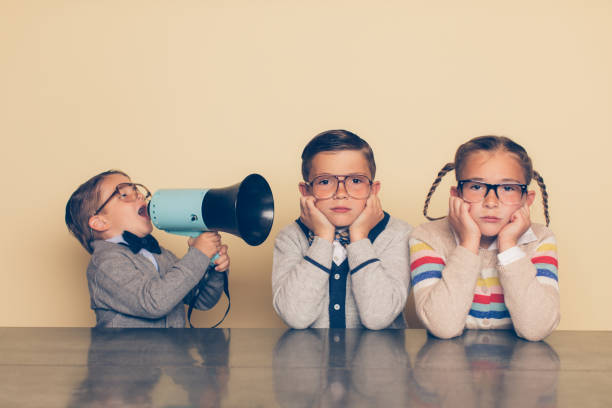 Young Nerd Boy Yelling at Siblings with Megaphone A young boy in glasses and a nerd outfit is shouting at his siblings through a megaphone. The big brother and big sister are looking at the camera with an annoyed look on their faces while plugging their ears. The little brother is bossing his older siblings around. nerd kid stock pictures, royalty-free photos & images