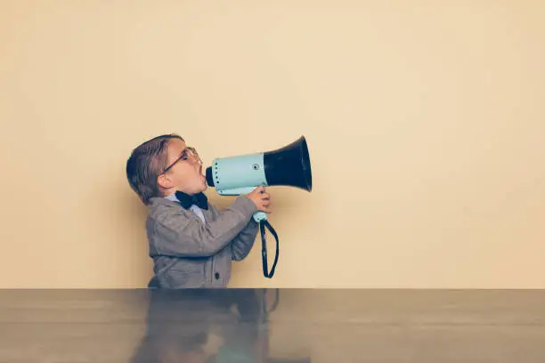 A young nerd boy yells into a megaphone in the studio. He is wearing a bow tie and eyeglasses. He wants you to listen to his important message.