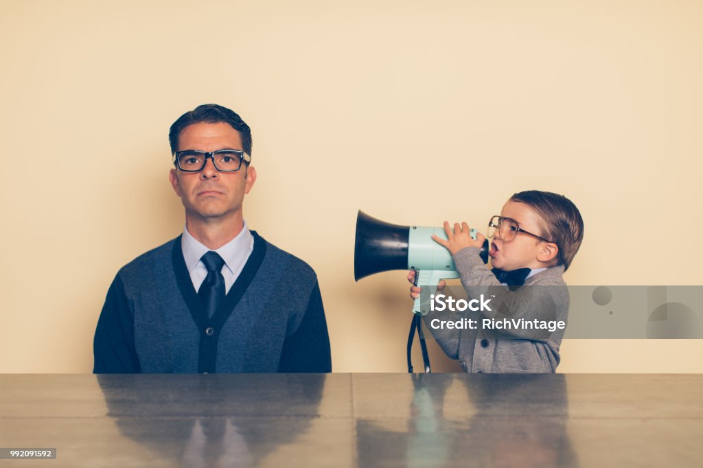 Young Nerd Boy Yelling at Dad through Megaphone A young nerd boy in eyeglasses and a sweater is yelling at his dad with a megaphone. His dad has a blank stare and is ignoring what the son is saying. The son is frustrated with his dad because he won't listen. Parents these days are distracted. Confusion Stock Photo