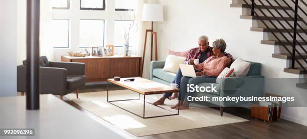 Foto de Casal Senior Sentado No Sofá Em Casa Usando O Laptop Para Fazer Compras Online e mais fotos de stock de Terceira idade