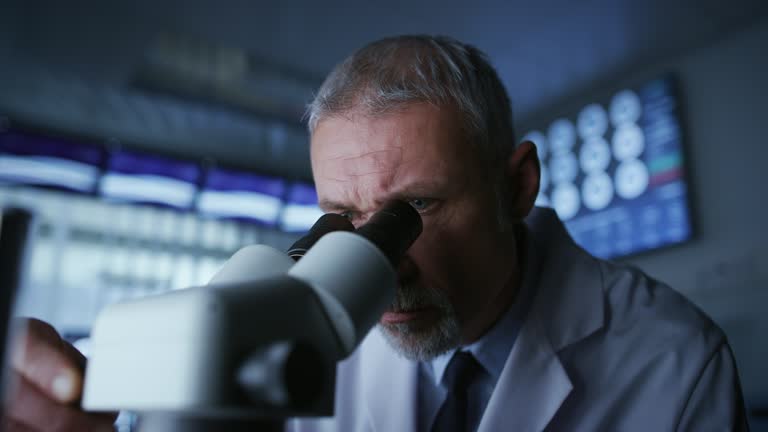 Senior Medical Research Scientist Looking under the Microscope in the Laboratory. Neurologist Solving Puzzles of the Mind and Brain. In the Laboratory with Multiple Screens Showing MRI / CT Brain Scan Images.