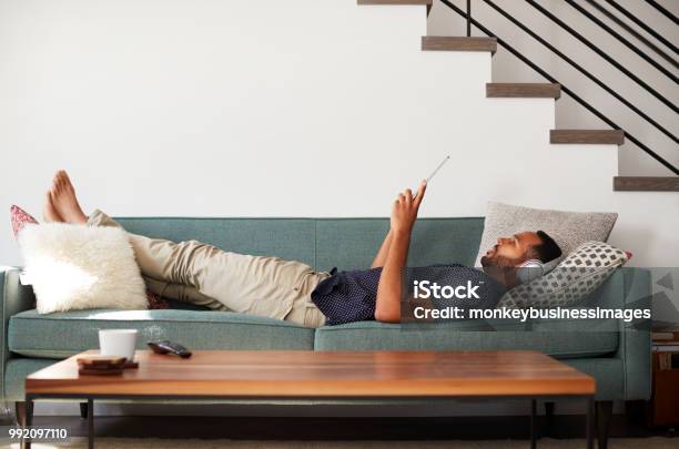 Man Lying On Sofa At Home Wearing Headphones And Watching Movie On Digital Tablet Stock Photo - Download Image Now