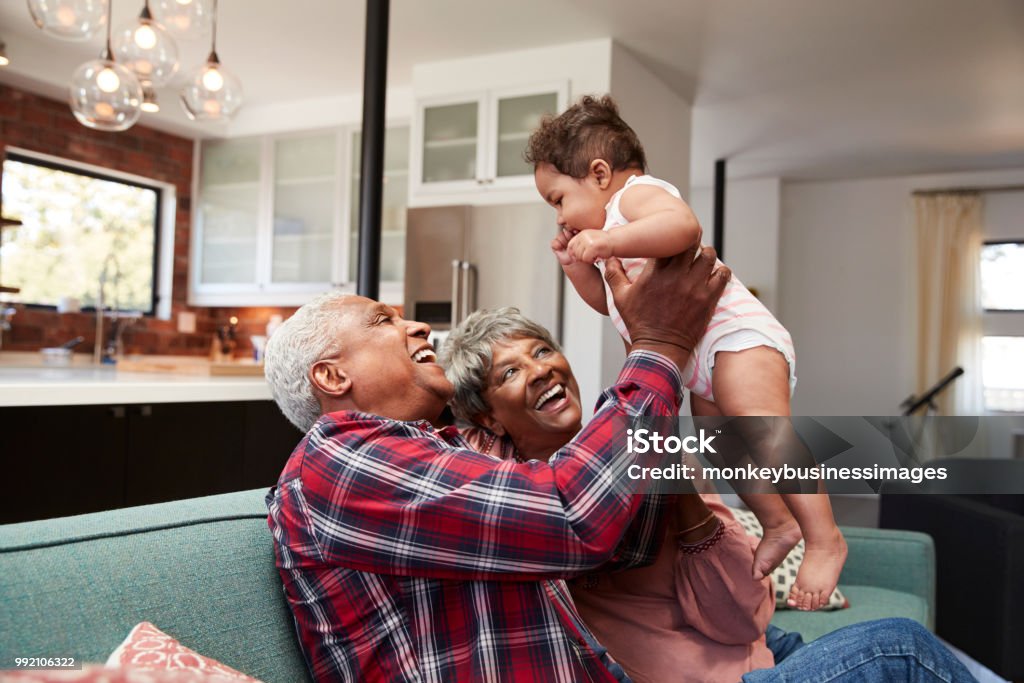 Grandparents Sitting On Sofa Playing With Baby Granddaughter At Home Grandparent Stock Photo